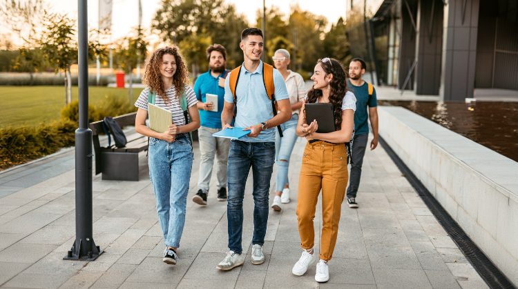 A group of smiling students walking outdoors on a college campus, carrying books and backpacks, enjoying a sunny day.